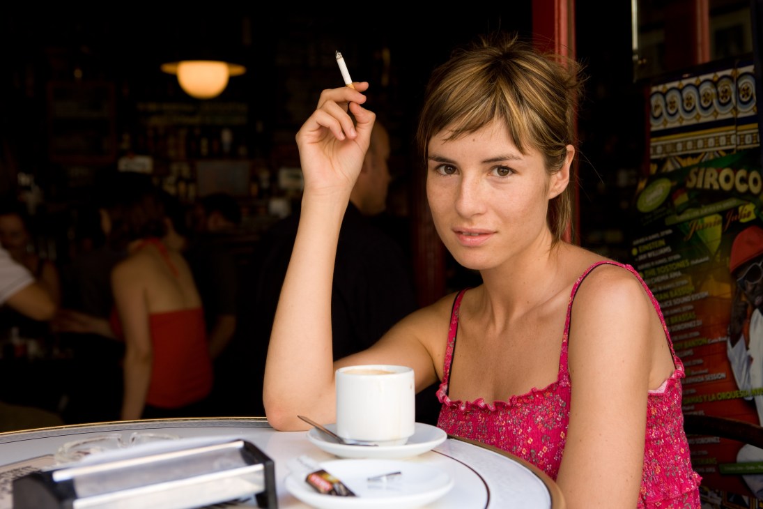 Patricia Jordi, an actress, sits at a café table holding a cigarette with a coffee cup in front of her.
