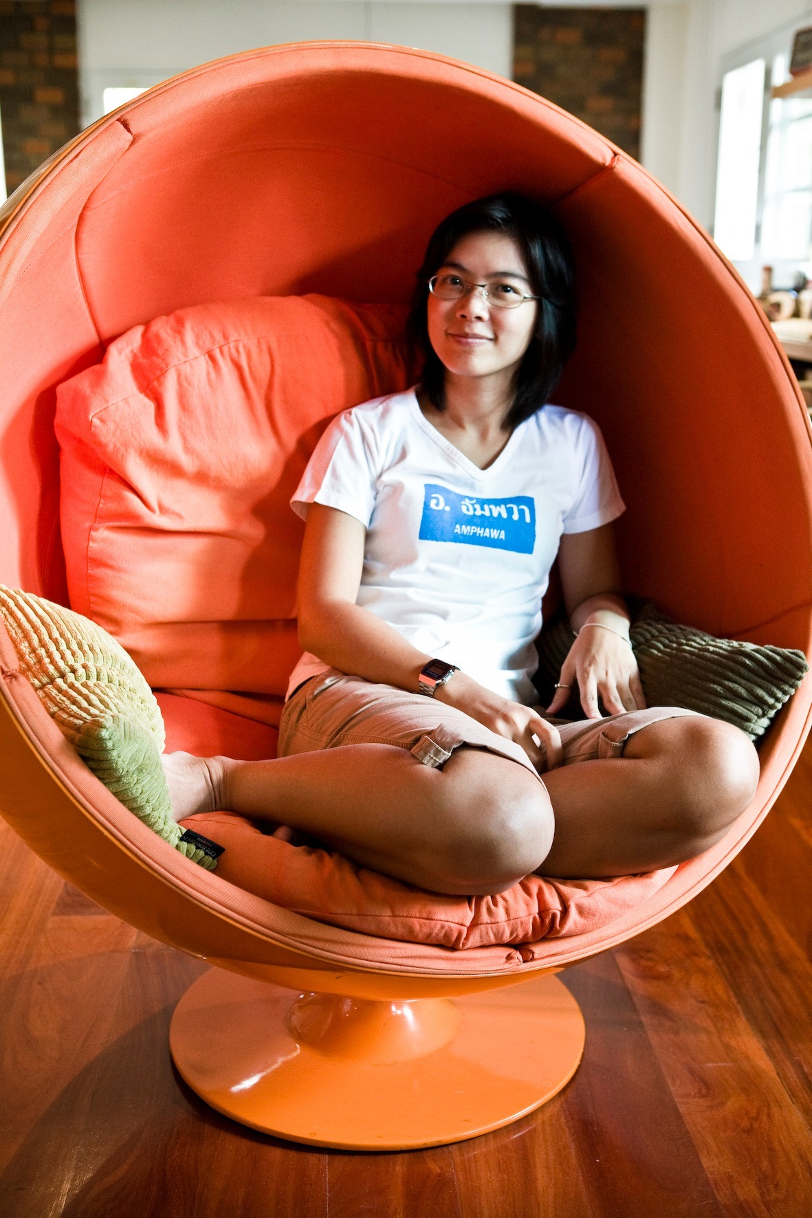 Woman sitting in a large orange egg-shaped chair, wearing glasses and a white t-shirt with blue text.