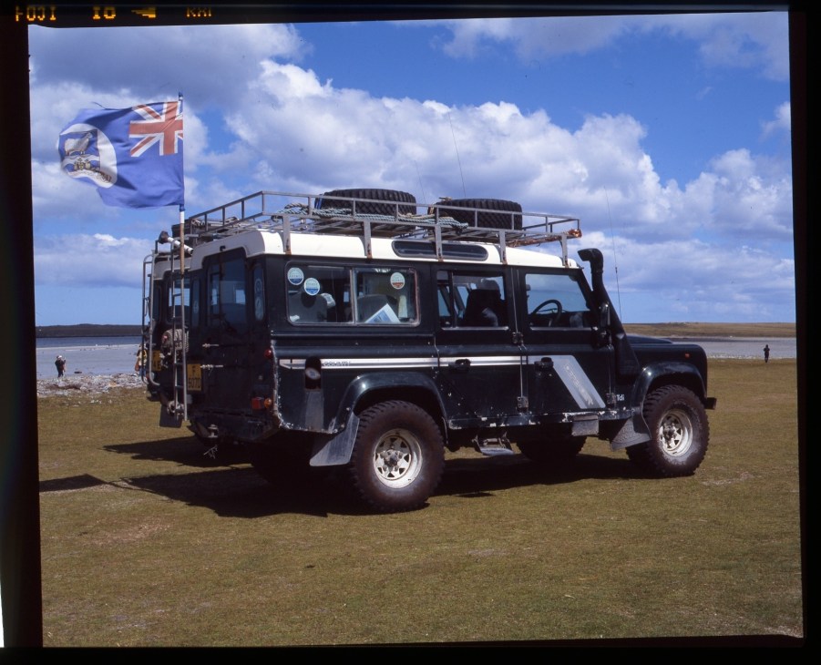 Black Land Rover with roof rack and Falkland Islands flag on a coastal beach.