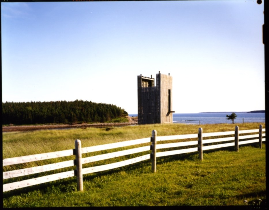 Ghost 6 tower, a tall black and natural wood modernist structure on a grassy coastal landscape with white fence in foreground, ocean and forest beyond.