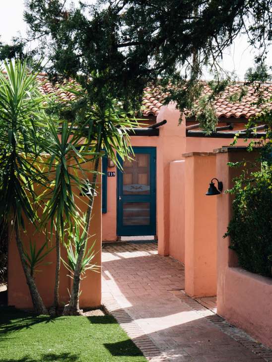 Coral-pink Spanish Colonial building with blue door at the Arizona Inn, framed by palm trees and desert vegetation.