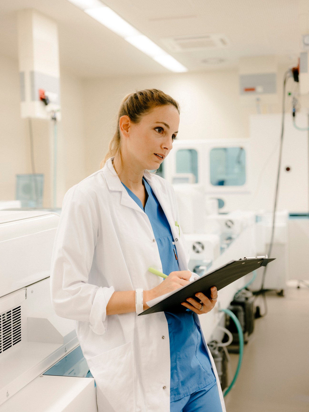 Female healthcare professional in white coat and blue scrubs holding a clipboard in a modern hospital laboratory.
