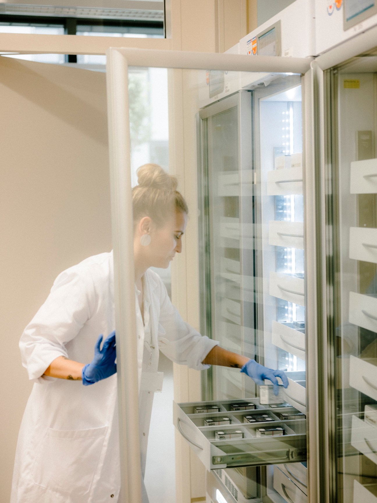 Female laboratory technician in white coat and blue gloves opening a refrigerated storage unit containing medical samples at Klinik Floridsdorf.