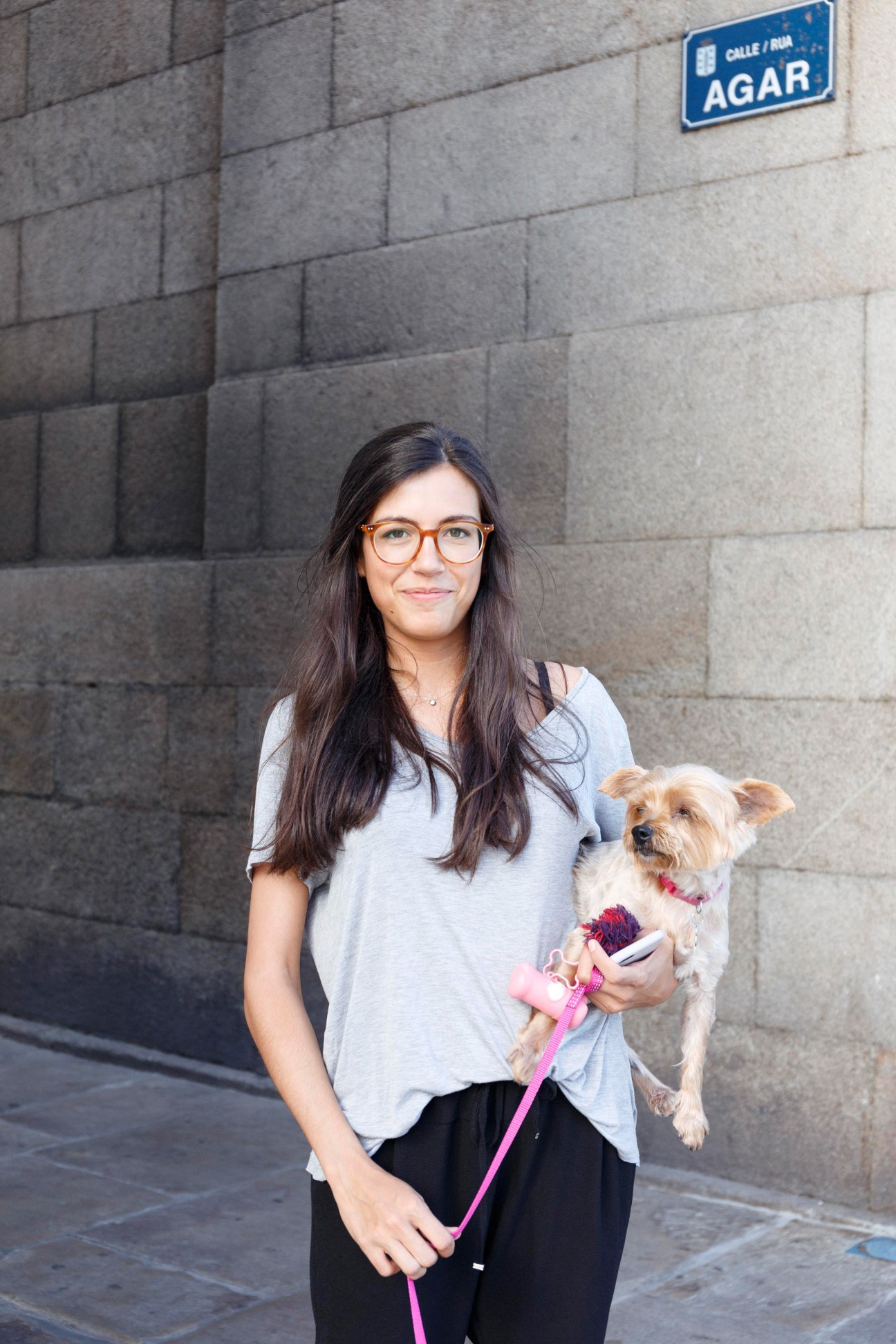 Woman with glasses holding a small tan dog on a city street with stone wall and street sign.