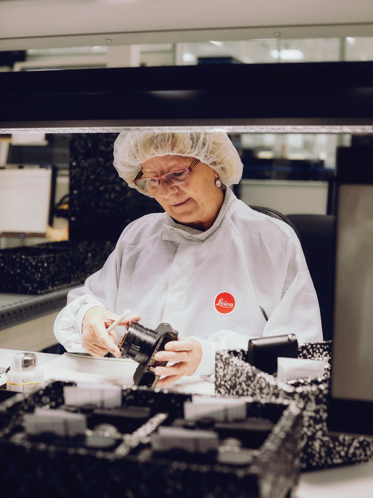 A Leica worker in a white lab coat and hairnet assembles a camera component by hand in a clean room.