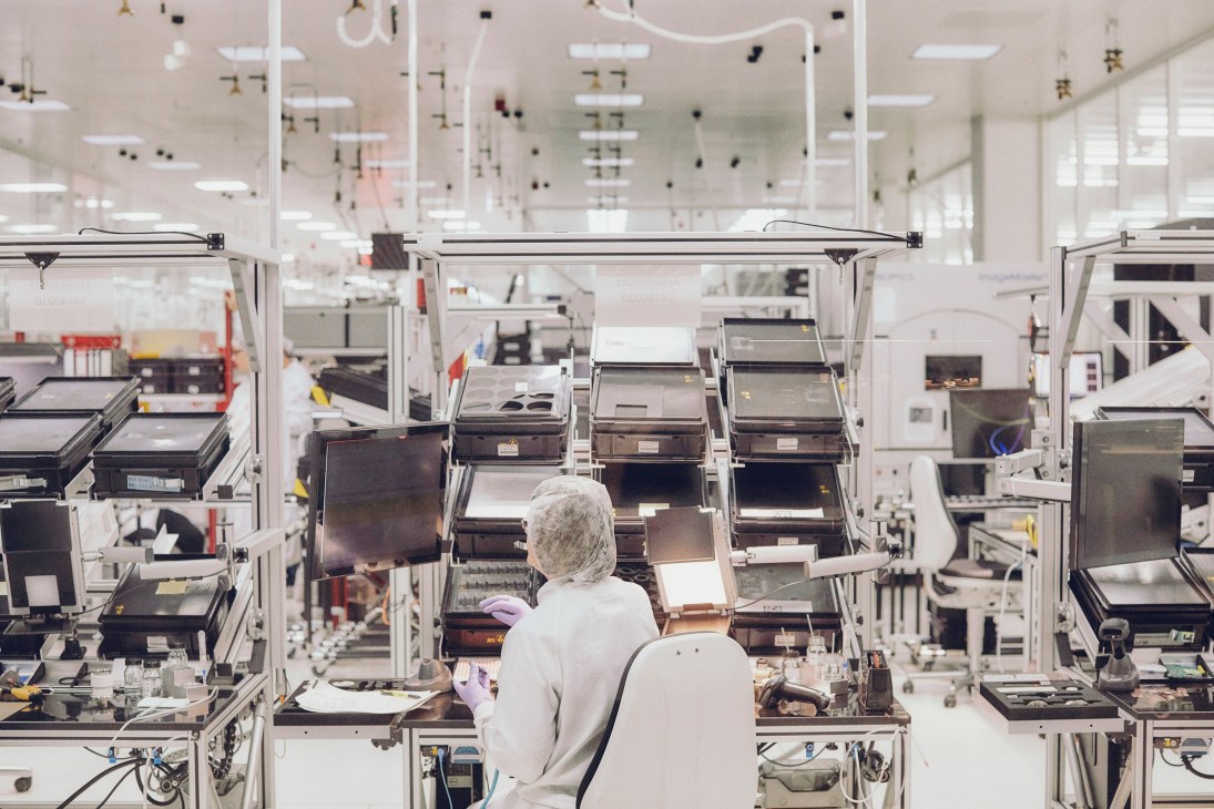 A worker in white lab coat and hairnet assembles camera lenses at workstations in Leica's clean room facility.