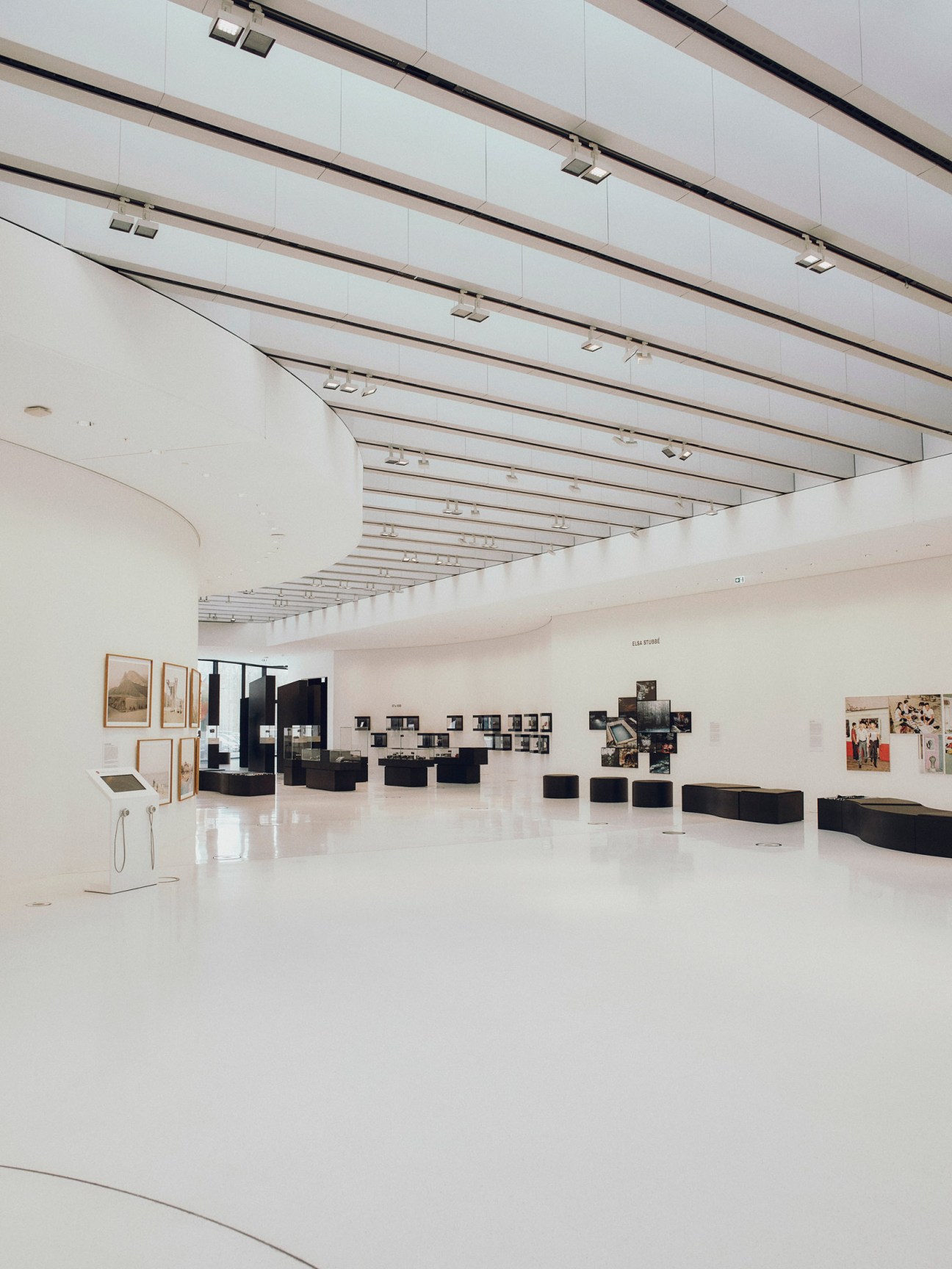 Minimalist gallery space with white walls, linear ceiling fixtures, and framed photographs displayed on walls and black benches.