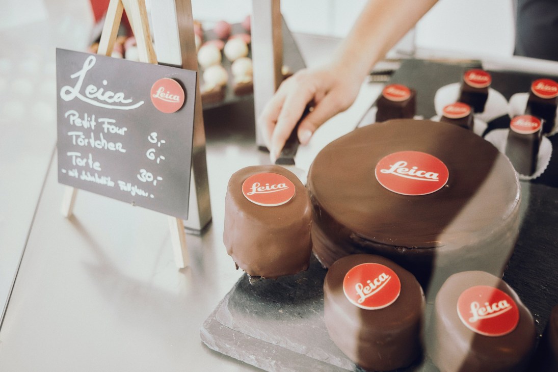 Chocolate desserts decorated with red Leica logos displayed on a counter with a menu board.