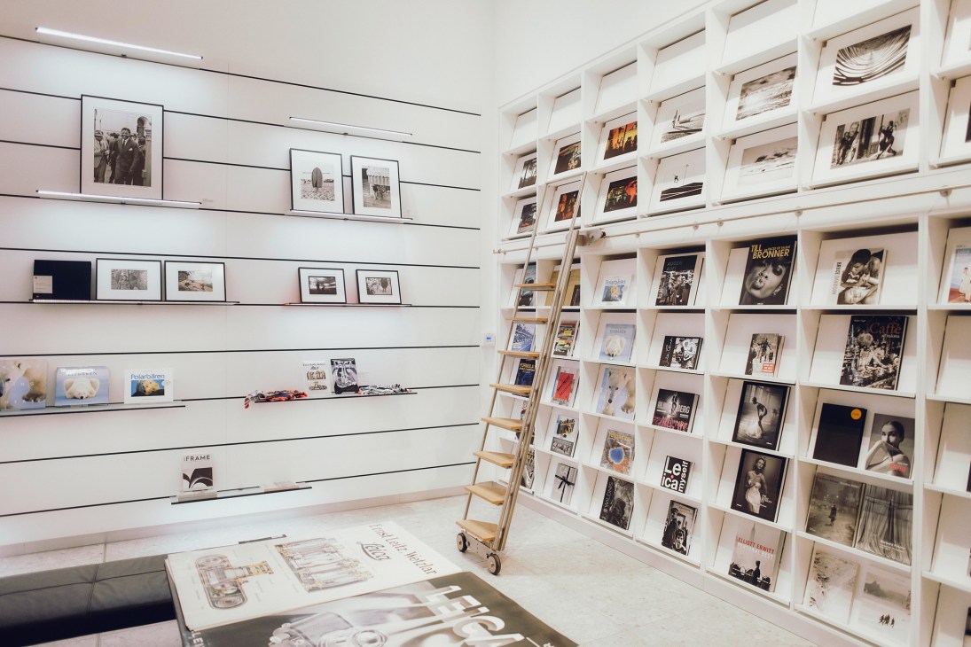 Interior of Leica museum shop displaying framed photographs and books on white shelving with a wooden ladder.