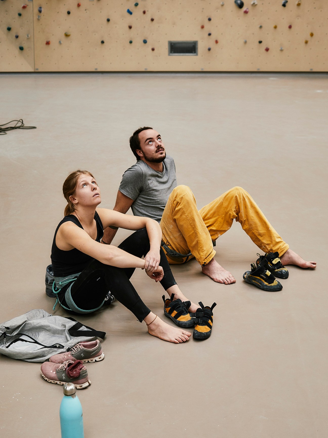 Two people sitting on floor of climbing gym looking up at climbing wall with colorful holds