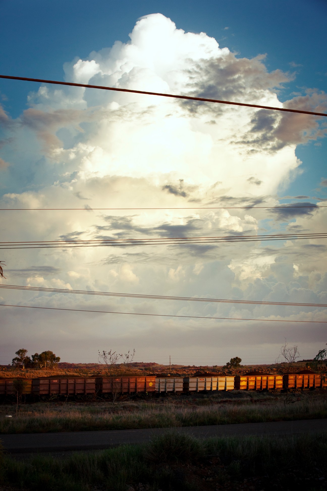 A freight train carrying mining products crosses the red Australian outback landscape under dramatic storm clouds.