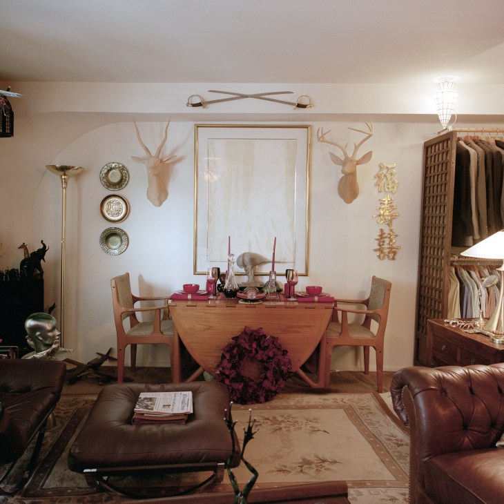 A studio apartment interior filled with collected objects, decorative deer heads, plates, furniture and clothing arranged throughout the space.