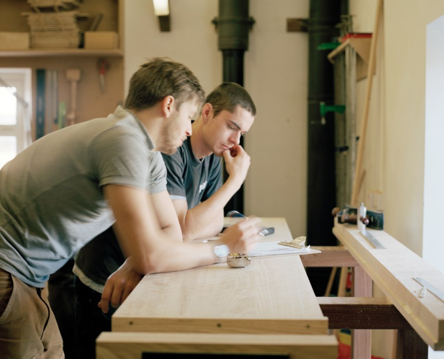 Two craftsmen examine woodwork plans at a workbench in a carpentry studio.