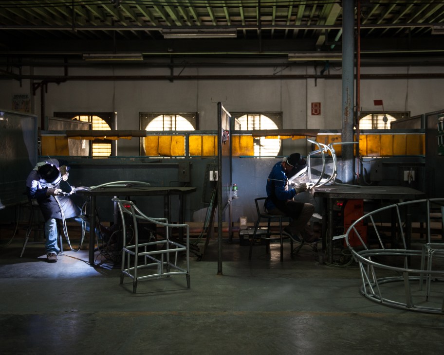 Workers welding metal furniture frames in an industrial factory production area with large windows.