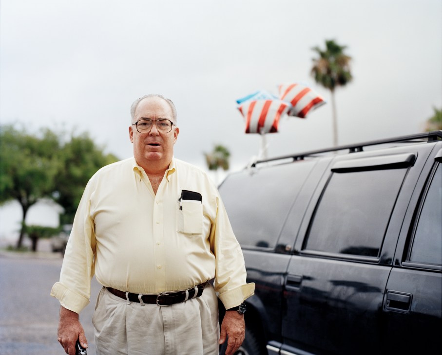 Man in yellow shirt standing in front of SUV with American flags and palm trees in background.