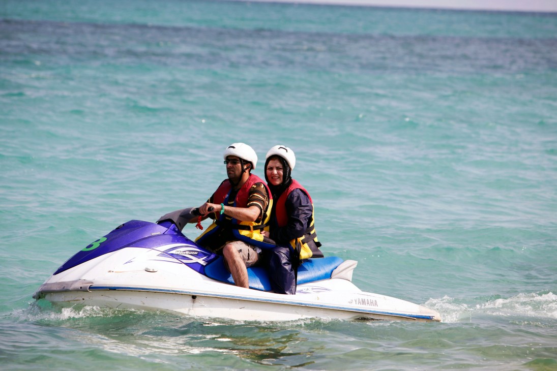 Two people on a jet ski in turquoise waters, both wearing white helmets and life jackets, with the woman wearing a hijab.