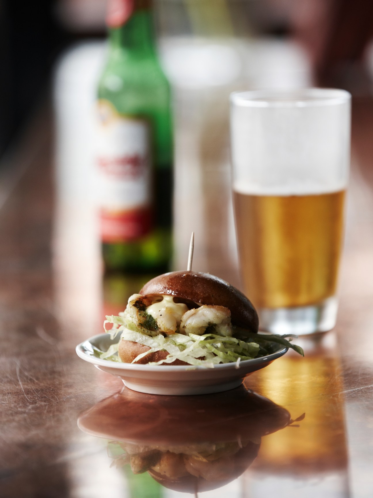 A burger with coleslaw on a plate next to a glass of beer and wine bottle.