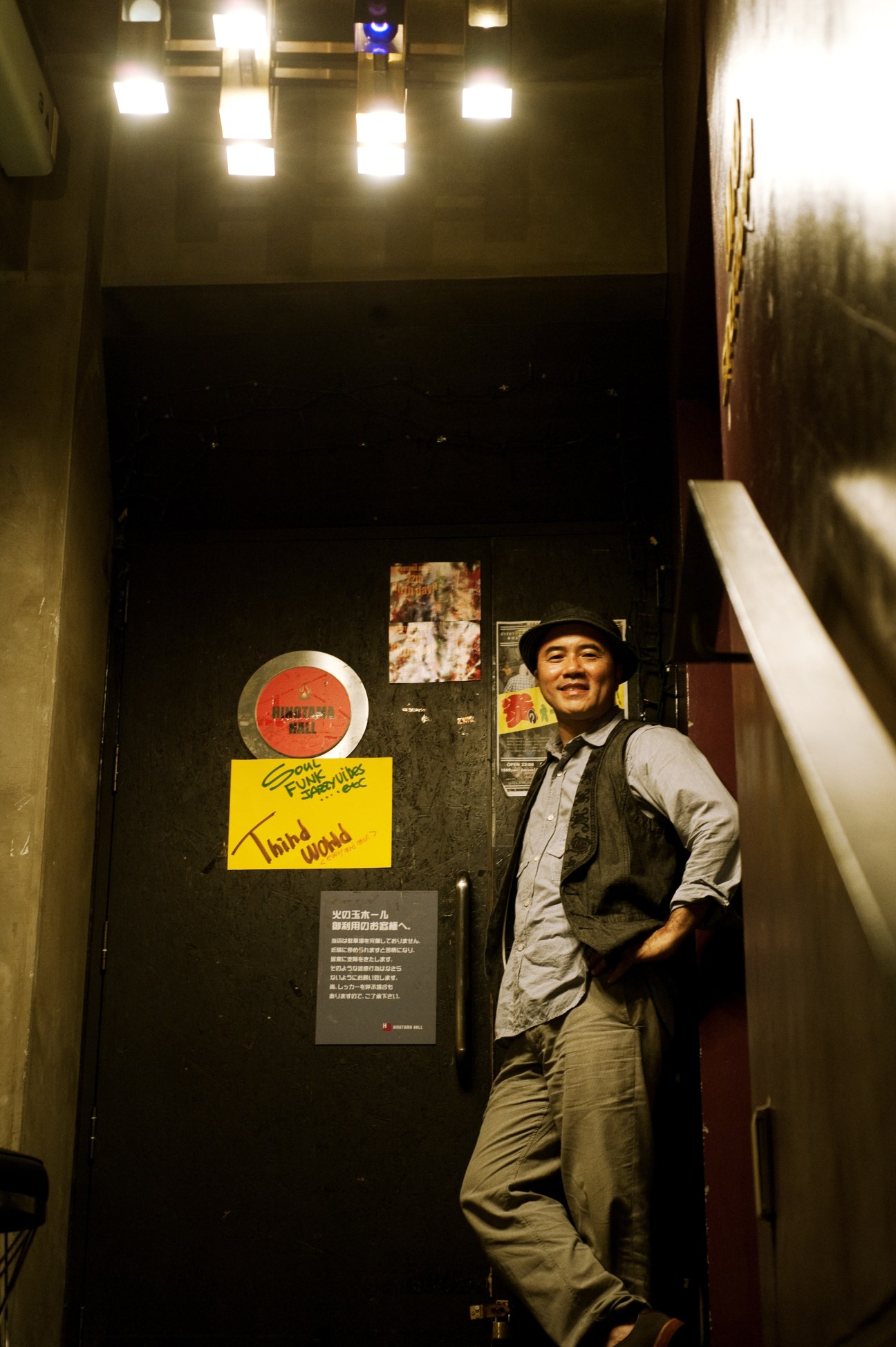 Kiichi, owner of night club Hinotama hall, leaning against a dark door decorated with stickers and signage.