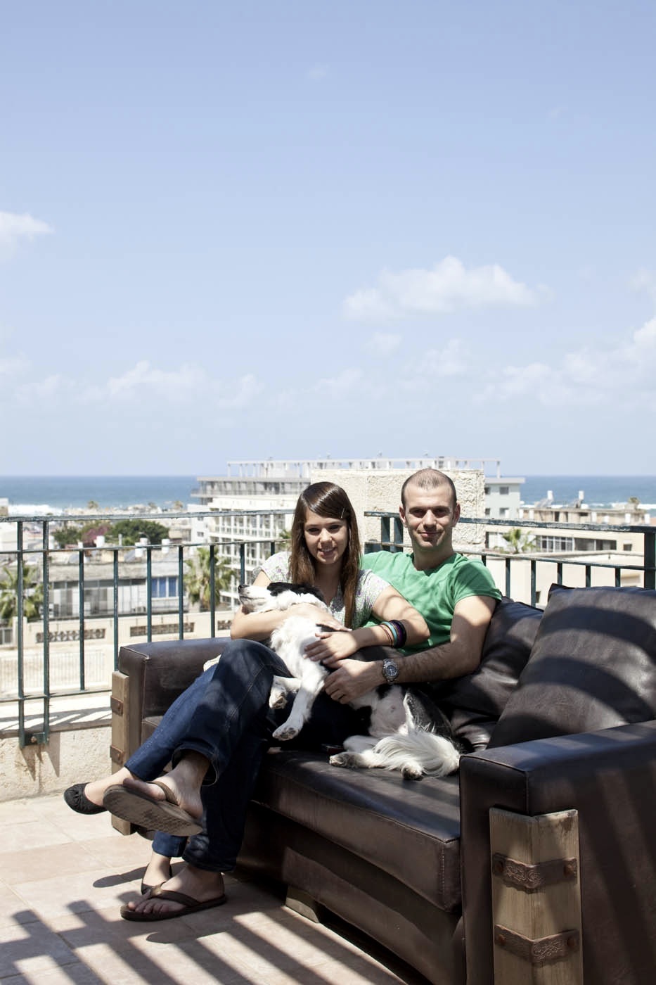 Ilan Blum and Ella Layani sit on a terrace sofa holding a small white dog, with Mediterranean Sea and Tel Aviv buildings visible behind them.