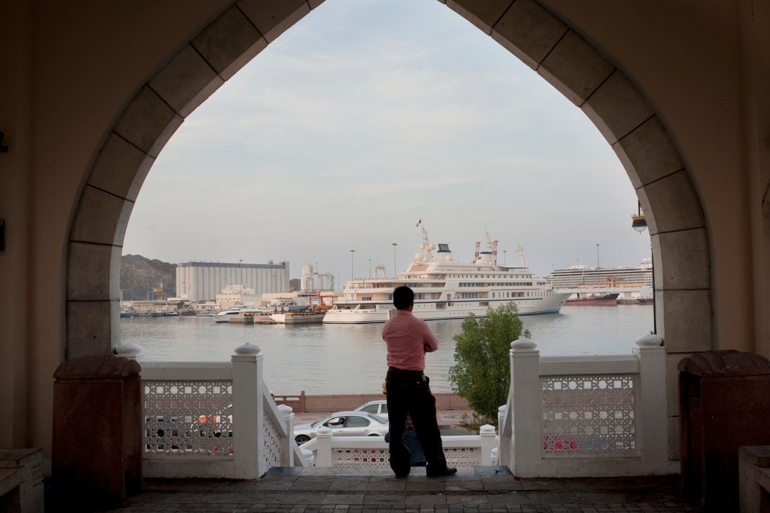 A person in a red shirt views a large cruise ship docked in Muscat harbour, framed by an ornate arched gateway.