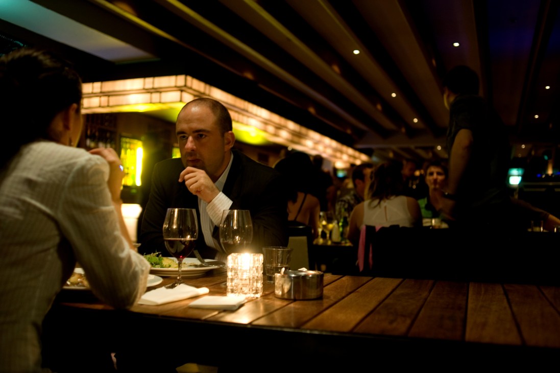 Man in dark suit dining at an upscale restaurant bar with wine glasses and warm lighting.