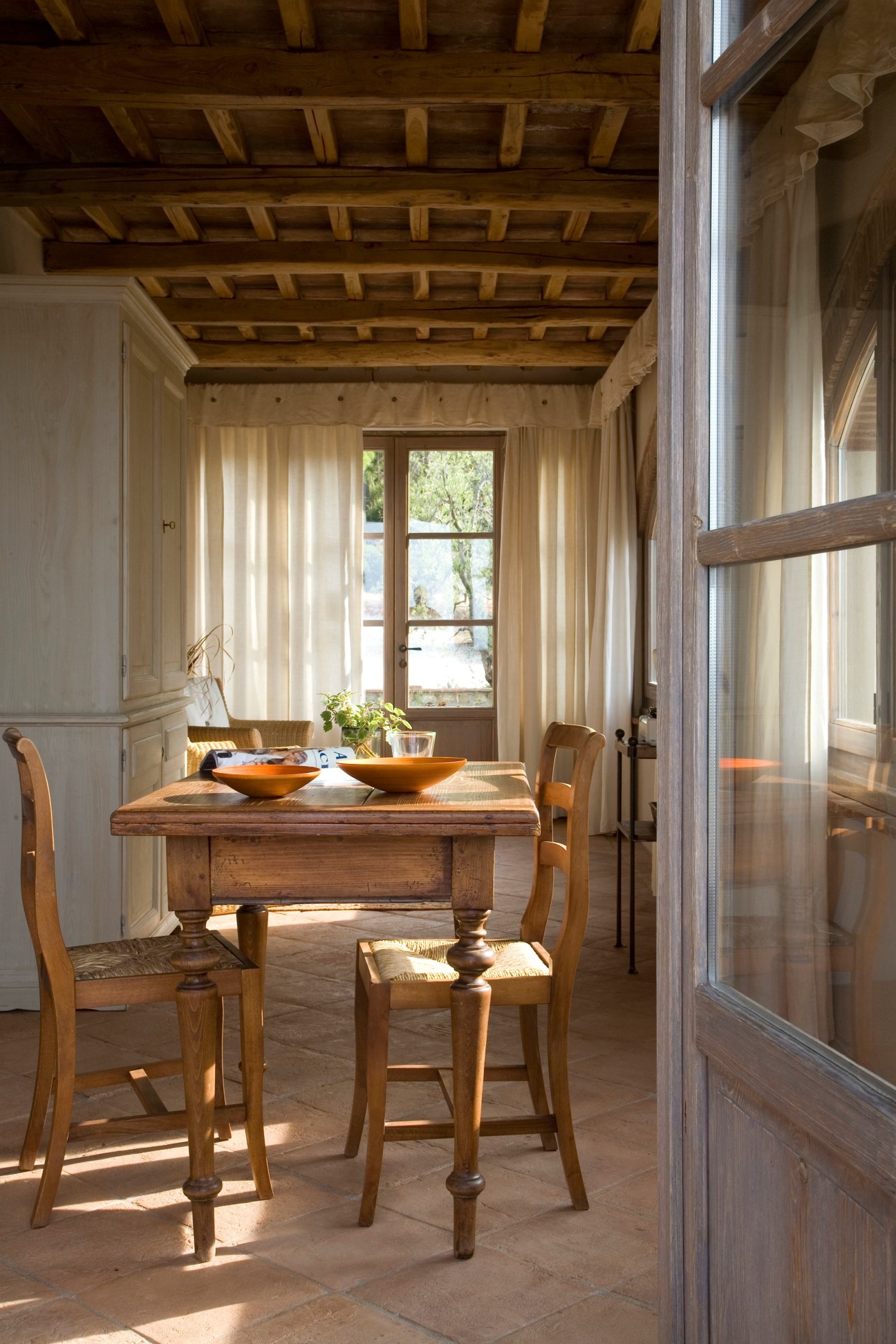 Sunlit dining room with wooden beamed ceiling, rustic table, chairs, and terracotta floor at Poggio ai Santi.