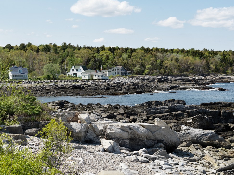 Rocky coastline with white clapboard houses and forested hills across blue water.