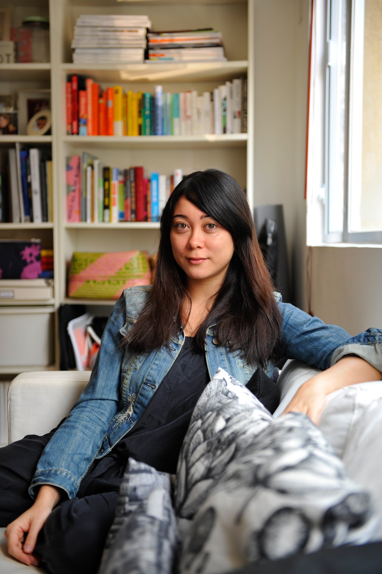 Danielle Huthart, a designer, sits on a couch in front of a white bookshelf filled with colorful books.