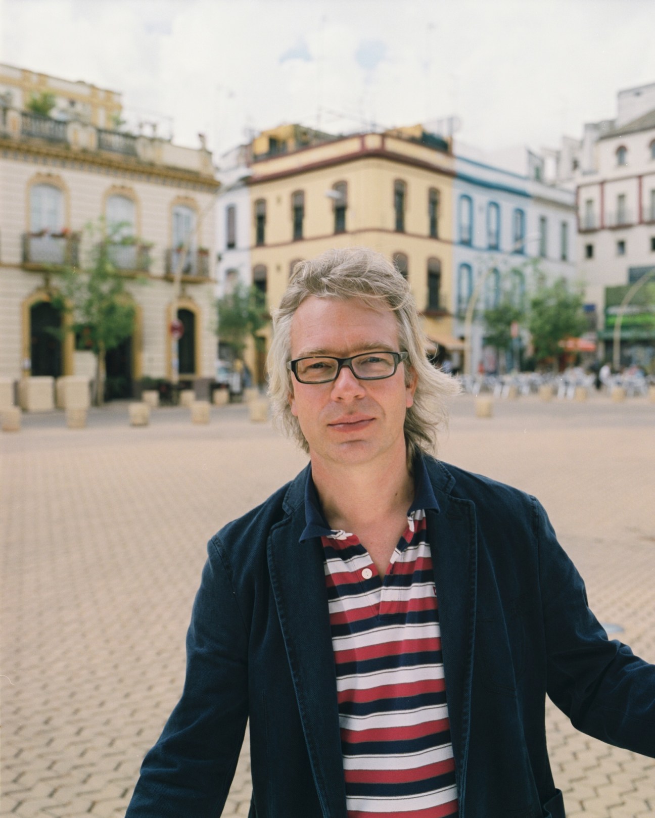 Man with grey hair and glasses standing in a plaza with historic buildings behind him.
