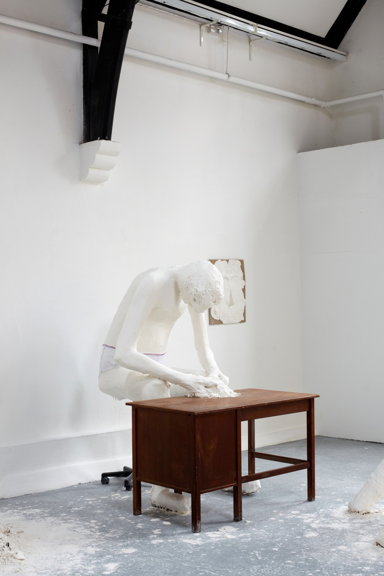 White plaster sculpture of a seated figure on a wooden desk in a gallery space.