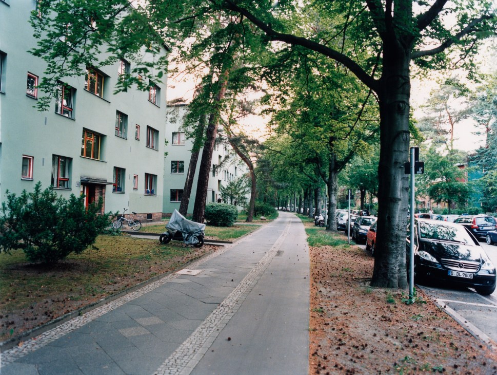 Tree-lined street with modernist apartment buildings and parked cars in a leafy Berlin suburb.