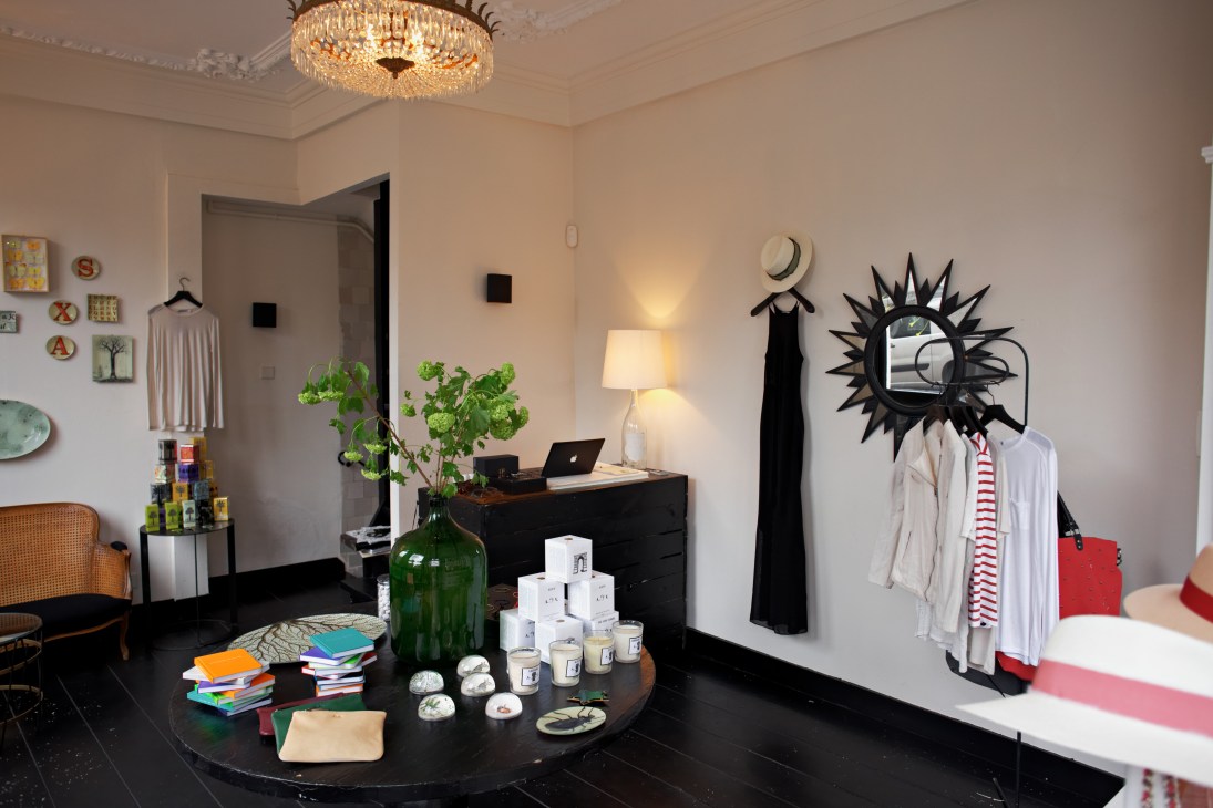 Maison Rika guesthouse interior showing concept boutique with clothing on display, decorative mirror, plants, and books on black round table.