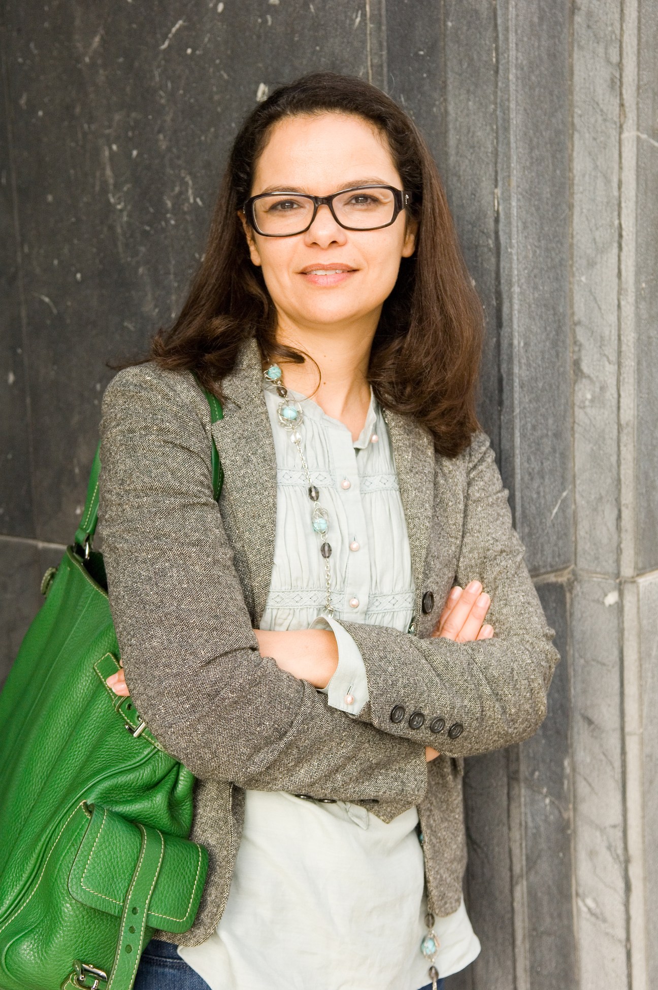 Sofia Alami, events manager, wearing glasses and a grey blazer with a green shoulder bag, standing against a dark textured wall.