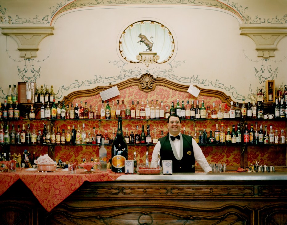 Bartender in formal attire stands behind an ornate bar with extensive spirits collection and baroque ceiling decoration.