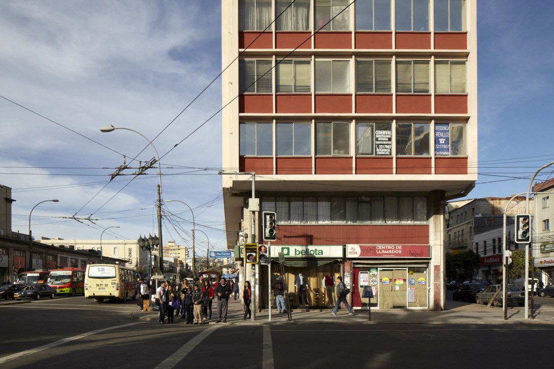 Street scene on Avenida Pedro Montt in Valparaíso showing a mid-century apartment building with red and cream facade, ground-floor shops, pedestrians, and a yellow bus.