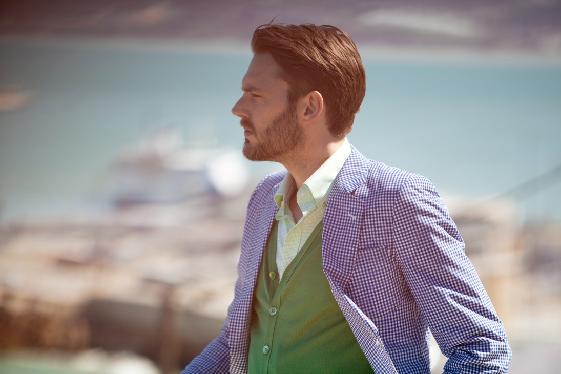 Man wearing purple checked jacket, green cardigan and white shirt posed against coastal backdrop.