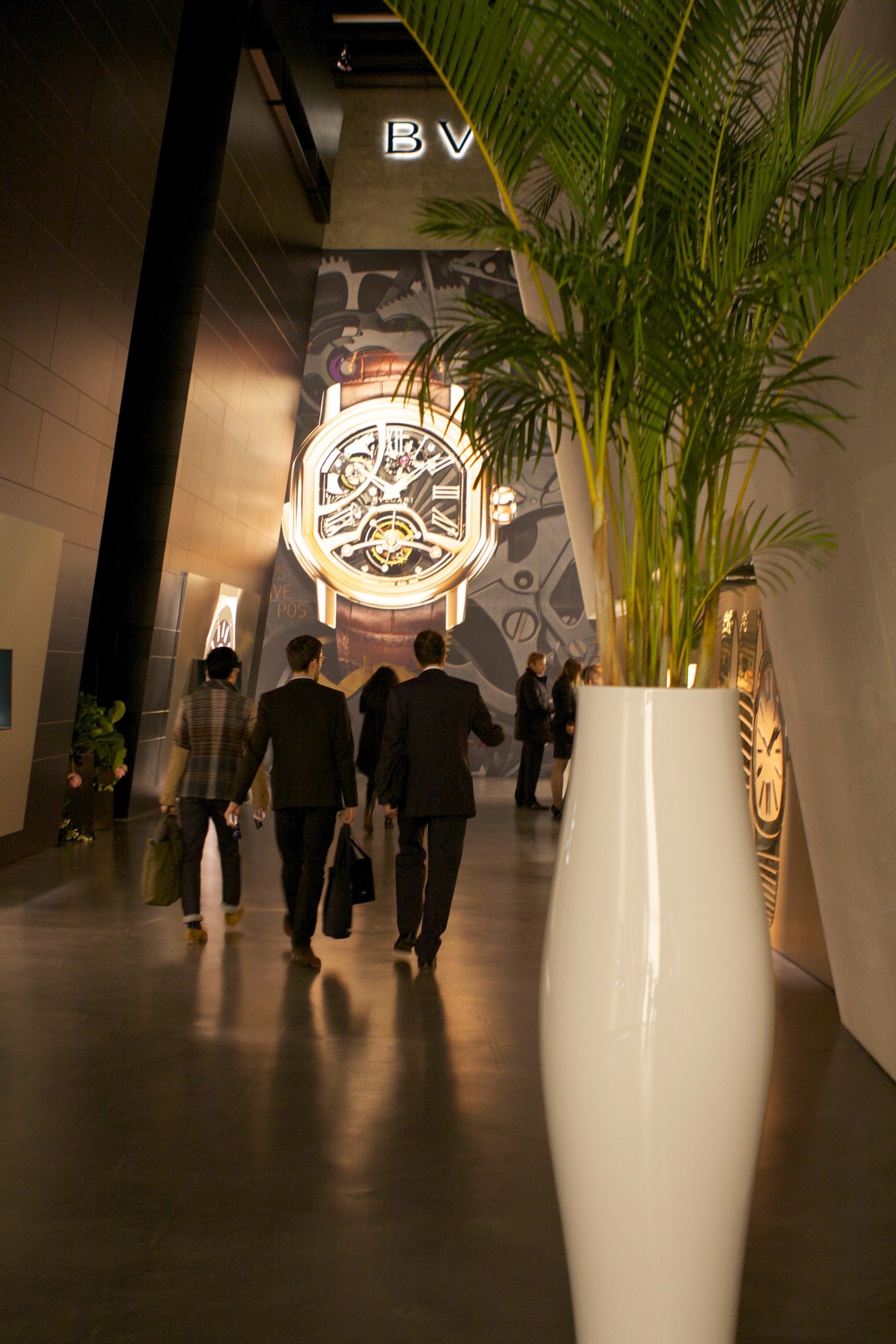 People walking through a modern watch fair exhibition hall with illuminated watch displays and green plant decorations.