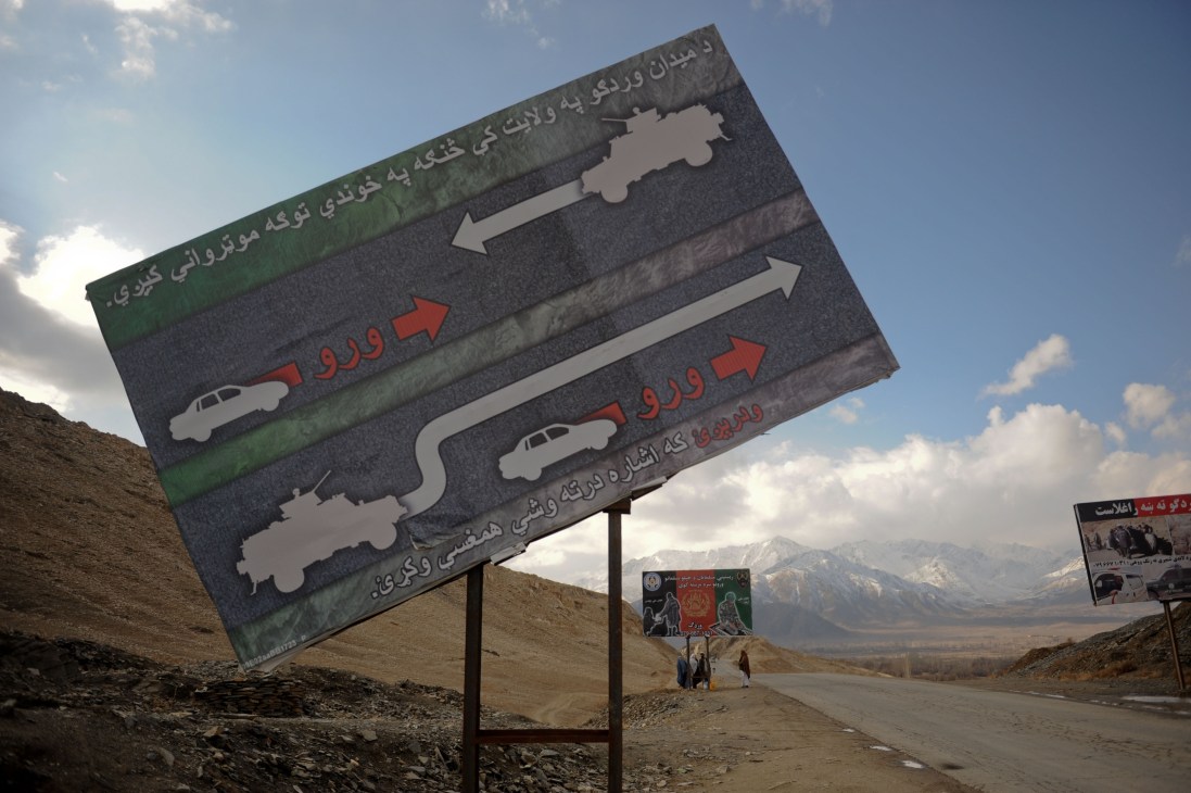 ISAF billboard with Dari and Pashtu text warning civilians to avoid military convoys, featuring vehicle silhouettes and arrows on Highway One in Wardak province.