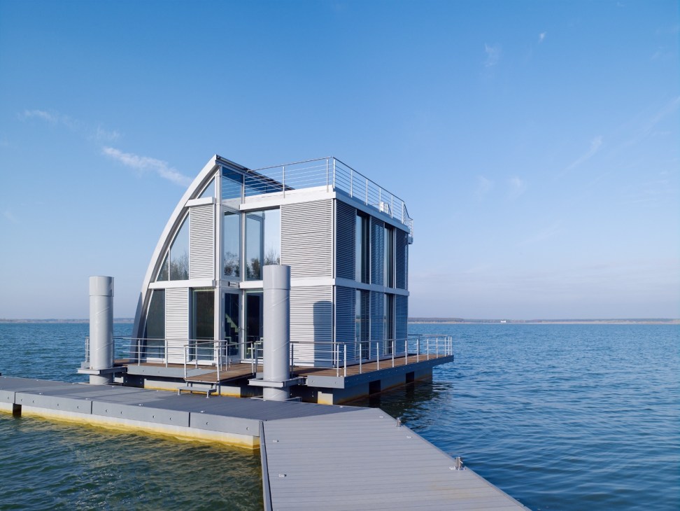 A modern floating home with white slatted exterior and glass windows sits on a platform in a calm blue lake.