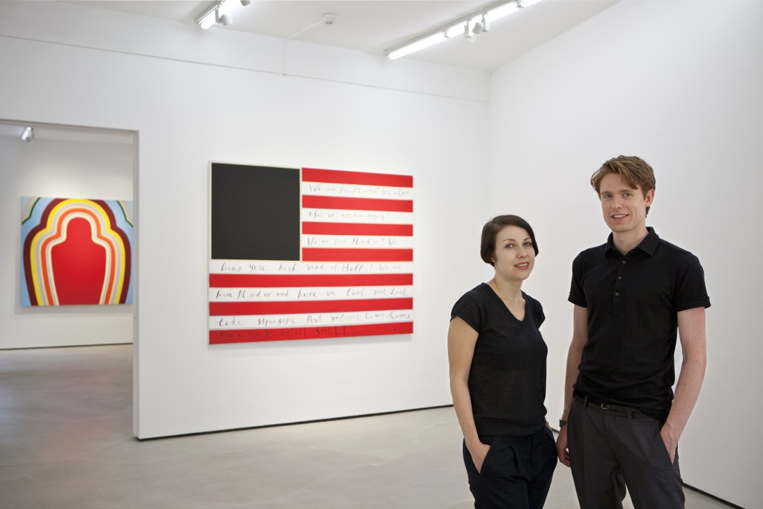Two gallery directors stand in a white gallery space with contemporary artworks on the walls behind them.