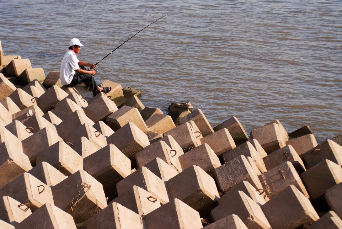 Fisherman casting a line from newly installed concrete flood barriers along Phnom Penh's riverfront.