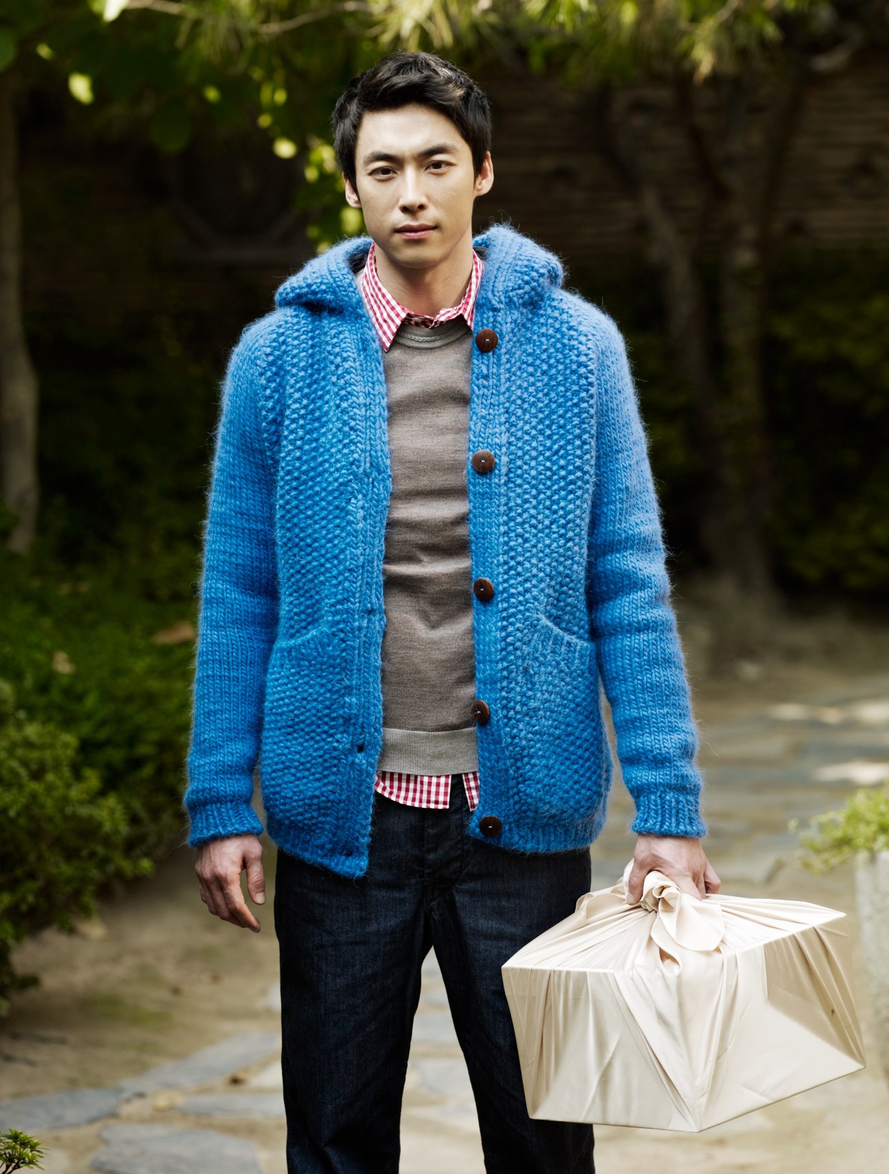 Man wearing blue cardigan, grey jumper, and checkered shirt holds a white umbrella outdoors.