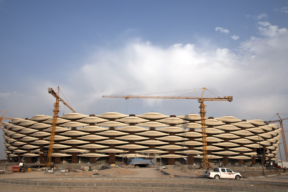 Construction site of Basra Sports City stadium showing distinctive woven pattern roof design with yellow cranes against blue sky.