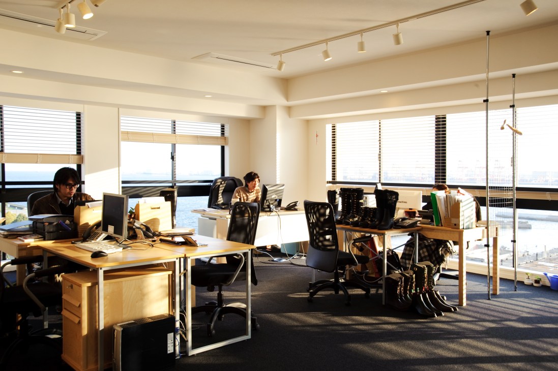 Bright office space with wooden desks, workers, and expansive windows overlooking Tokyo Bay and waterfront views.