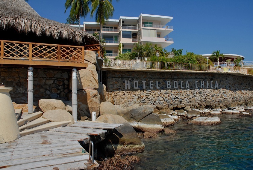 Boca Chica Hotel in Acapulco with stone facade, wooden dock, and modern white building perched above rocky coastline.