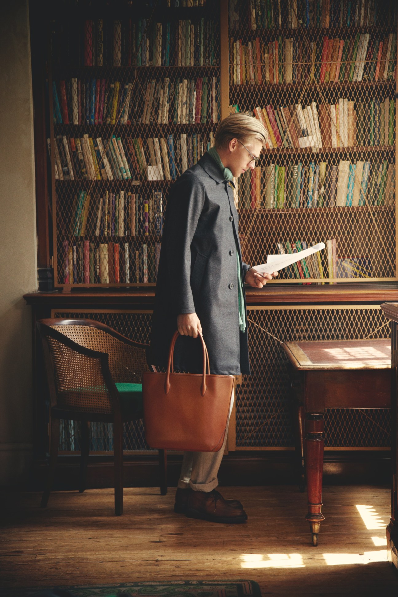 Person in grey coat and glasses holding a document and leather bag in a library setting