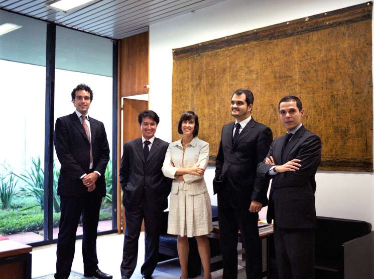 Five young diplomats in business formal attire stand in a modern ministry office with floor-to-ceiling windows.