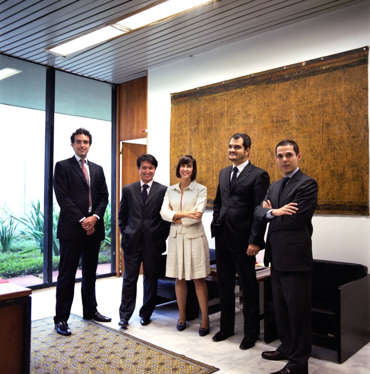 Five young diplomats in business formal attire stand in a modern ministry office with floor-to-ceiling windows.