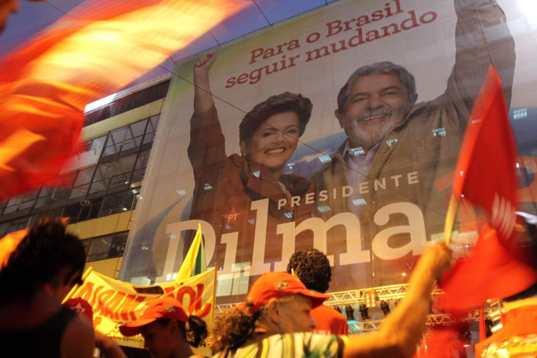 A large campaign billboard showing two smiling figures with Portuguese text, surrounded by supporters holding red flags at a nighttime rally.