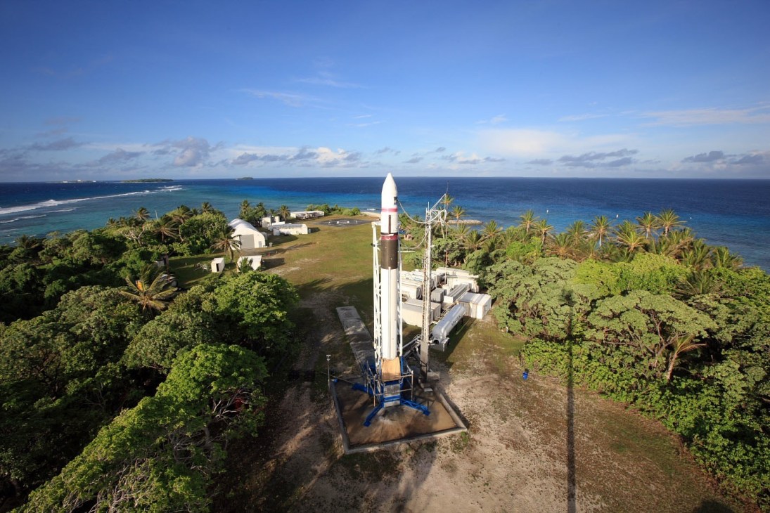 Aerial view of a white rocket on a launch pad surrounded by tropical vegetation and ocean.